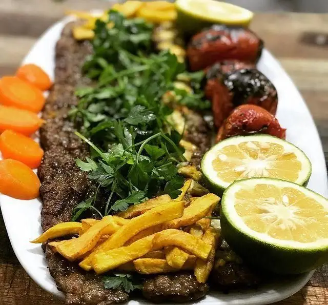 سینی کباب کوبیده و گوجه کبابی رستوران حمید با دورچین‌های جذاب شامل سیب‌زمینی، لیموترش، هویج و جعفری تازه. غذای لذیذ ایرانی.