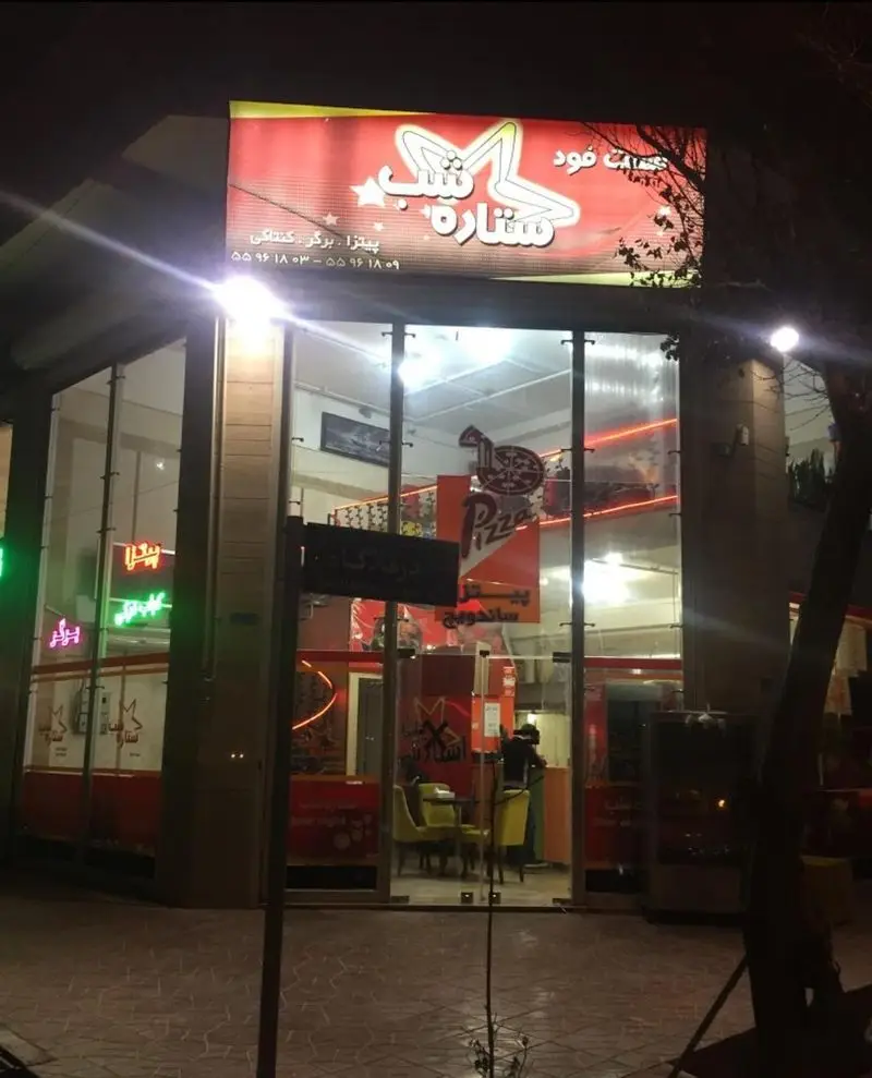 تصویری در شب از نمای بیرونی فست فود ستاره شب با تابلوی روشن، ورودی شیشه‌ای. ارائه پیتزا، برگر و ساندویچ خوشمزه.