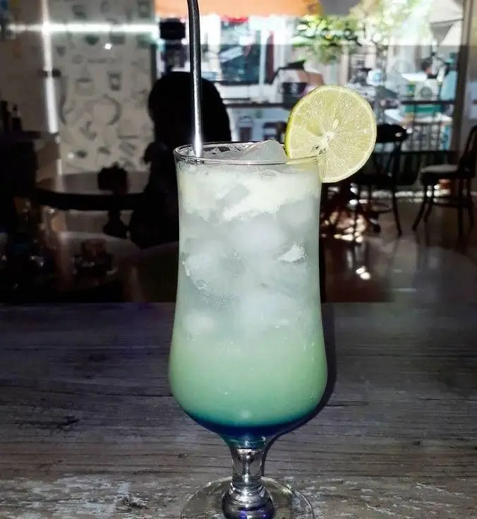 تجربه نوشیدنی خنک و دلپذیر در کافه فراز. کوکتل رنگی با لایه های آبی و سبز، یخ و برش لیمو، انتخابی عالی برای اوقات فراغت.