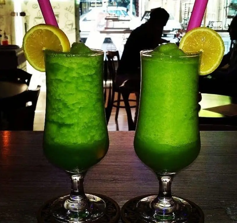 دو نوشیدنی سبز یخی و خنک با برش‌های لیمو و نی صورتی روی میز چوبی در محیط دنج کافه فراز. طعمی دلنشین و گوارا.