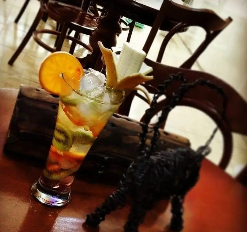 تصویری جذاب از نوشیدنی میوه‌ای خنک (کیوی، پرتقال) همراه با یخ در لیوانی بلند، سرو شده بر روی میز چوبی کافه فراز.