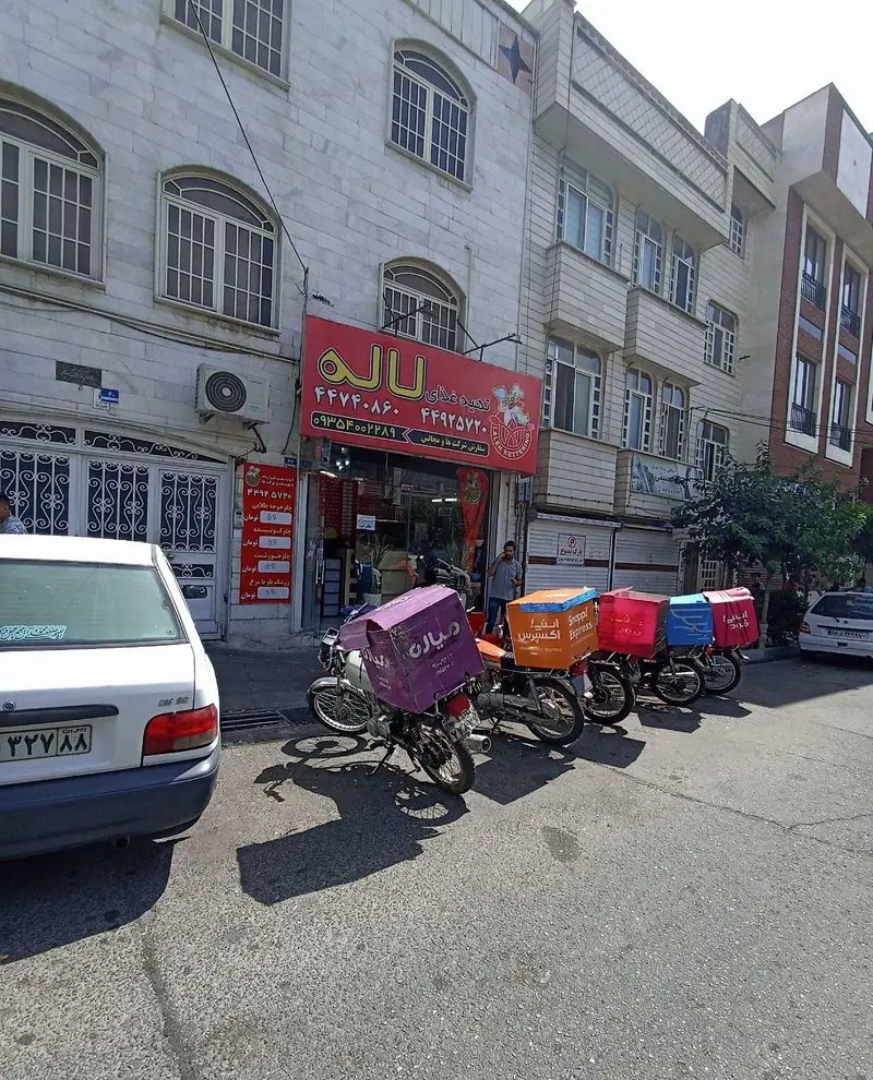 نمای بیرونی کترینگ و تهیه غذای لاله با تابلوی روشن و موتورسیکلت‌های دلیوری در خیابان تهران، آماده ارسال غذای ایرانی و مجلسی.