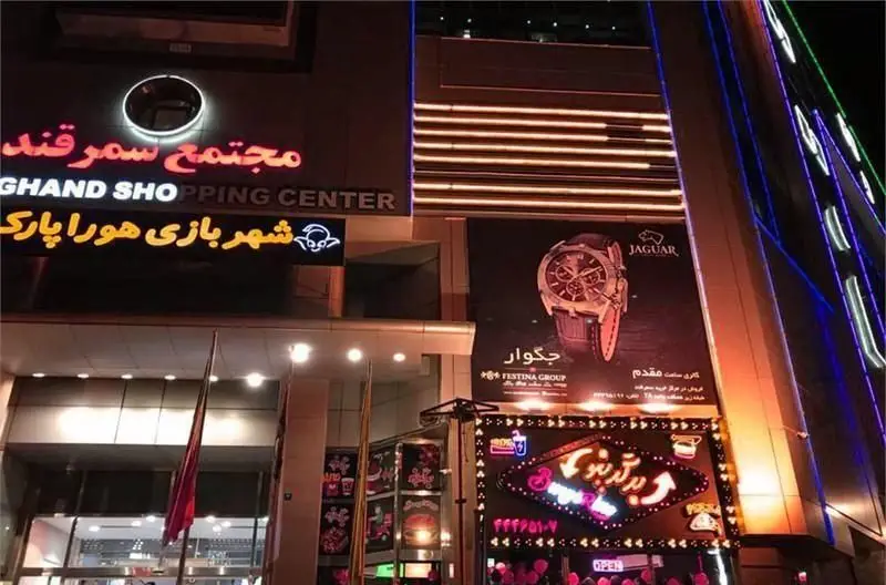 ورودی مجتمع سمرقند در شب با روشنایی خیره کننده، تابلوی نئون برگرینو، شهر بازی هورا پارک و بنر ساعت جگوار.