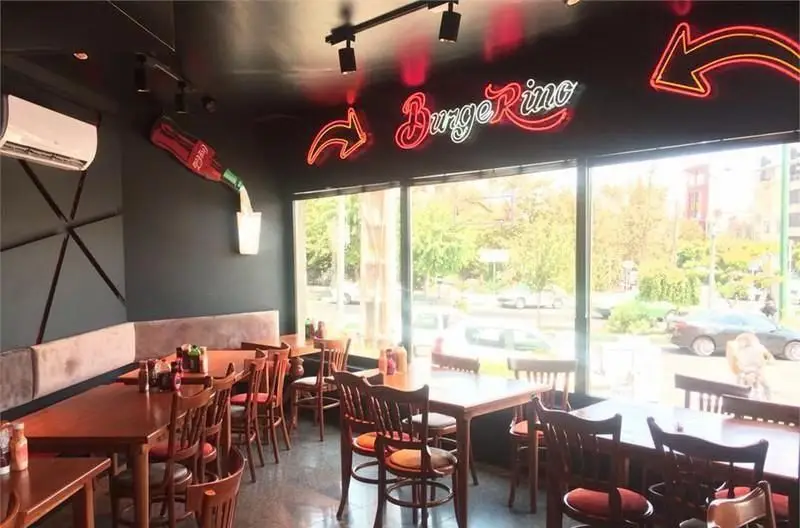 فضای داخلی رستوران مدرن برگرینو با میز و صندلی چوبی، نیمکت راحتی، پنجره‌های بزرگ و دکوراسیون خلاقانه بطری نوشابه و تابلو نئون.