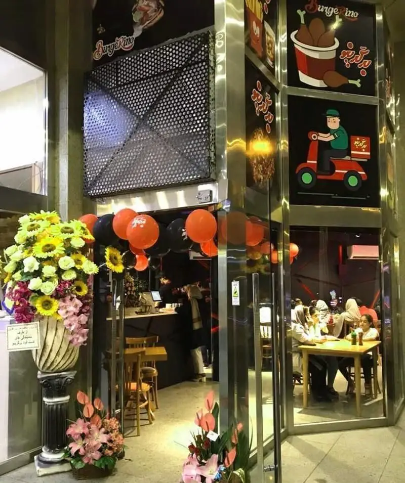 تصویری از نمای داخلی و بیرونی رستوران فست فود برگرینو، با دکوراسیون چشمگیر، مشتریان و تابلوهای تبلیغاتی غذاها از جمله همبرگر و مرغ سوخاری.