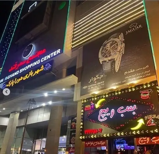 تصویر شبانه ورودی فست فود برگرینو با تابلو نئون جذاب "بمب و پیتزا" همراه با آیکون‌های برگر و پیتزا در مجتمع تجاری گرند.