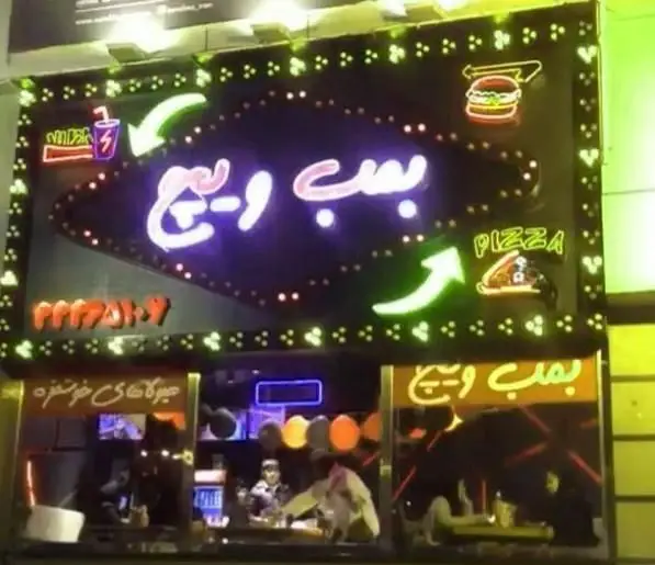 تصویری از تابلوی نئون فست فود برگرینو در شب با نام "بمب و پیتزا" و نمادهای برگر، پیتزا و نوشیدنی به همراه شماره تماس.