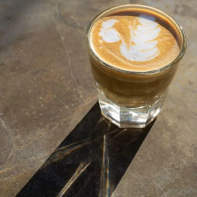قهوه لاته خوش طعم کافه رستوران لول ۴، با لاته آرت زیبا و سایه دلنشین آن زیر نور آفتاب بر میز سنگی.