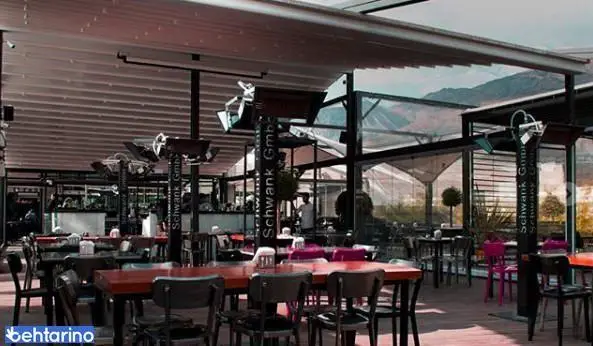 نمای داخلی کافه رستوران لول ۴ با فضای باز و سقف متحرک، میز و صندلی‌های چوبی، هیترهای Schwank و منظره زیبای کوهستانی.