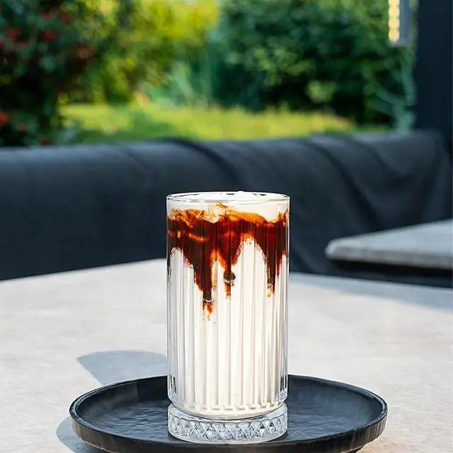 نوشیدنی لایه ای شکلاتی و شیری در لیوان شیاردار، روی میز کافه رستوران لول ۴، در فضای سبز دلنشین تابستانی.