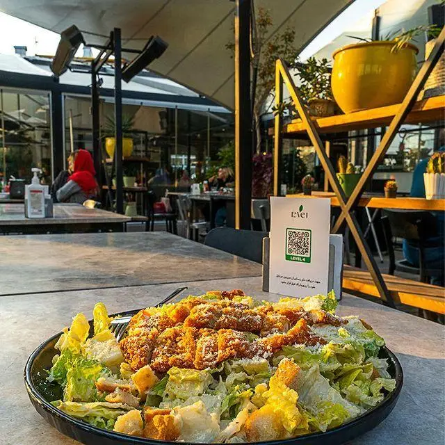 سالاد سزار خوش‌رنگ و لعاب با تکه‌های مرغ سوخاری در کافه رستوران لول ۴، به همراه منوی QR و فضای باز پر از مشتریان.