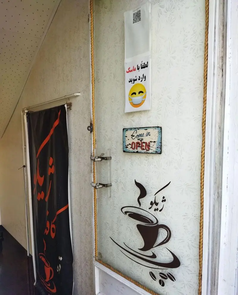 ورودی کافه ژیکو با درب شیشه‌ای مزین به طرح فنجان قهوه، تابلو "Come in we're OPEN" و اطلاعیه ورود با ماسک.