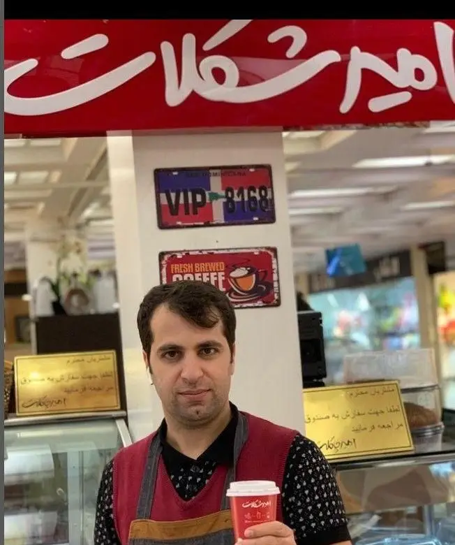 باریستا در کافی شاپ امیرشکلات، لیوان قهوه ای در دست دارد. نام برند امیرشکلات و تابلوی "Fresh Brewed Coffee" در پس زمینه دیده می‌شود.