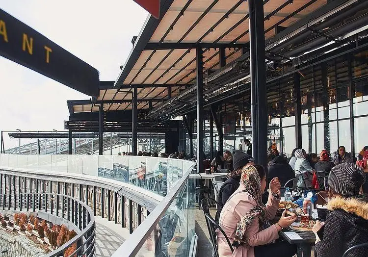 نمایی از فضای باز فود پارک فلای لند با سقف متحرک، تراس شیشه‌ای و مشتریان در حال صرف غذا. رستورانی با منظره عالی.