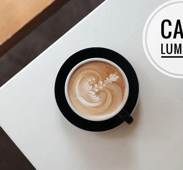 یک فنجان قهوه لاته آرت زیبا با طرح برگ روی میز سفید، نمایی جذاب از فضای کافه رستوران لومیر برای دوستداران قهوه و آرامش.