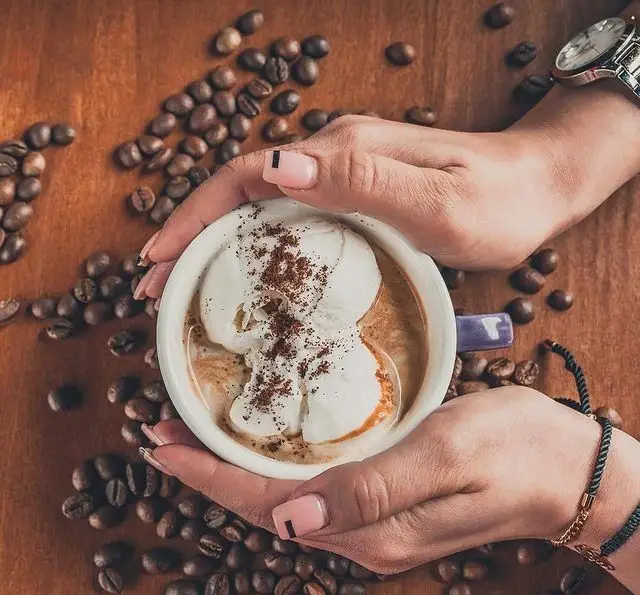 نمایی نزدیک از دستان لطیف، در حال نگهداری یک فنجان قهوه بستنی خوشمزه در کافه رستوران لومیر. دانه‌های قهوه در پس‌زمینه. فضایی گرم.