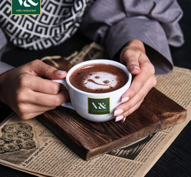 مشتری در کافه رستوران لومیر فنجانی از هات چاکلت (قهوه) با طرح لبخند را در دست دارد. روی تخته چوبی و روزنامه قرار گرفته.