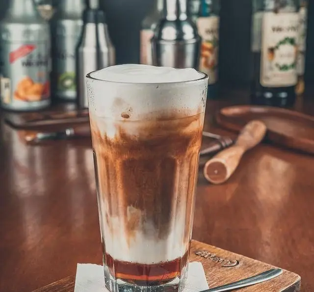 تصویری زیبا از یک لیوان بلند نوشیدنی سرد لایه‌ای قهوه با کف غلیظ سفید در کافه رستوران لومیر، آماده سرو برای میهمانان.