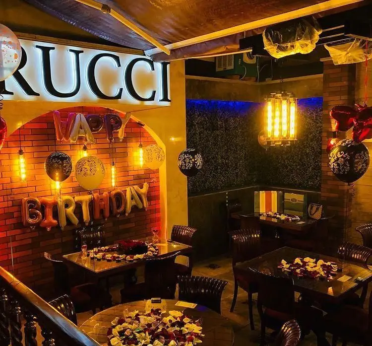 دکوراسیون شیک رستوران ایتالیایی روچی برای جشن تولد، با بادکنک‌های HAPPY BIRTHDAY، نورپردازی گرم و میزهای رمانتیک با گلبرگ گل.