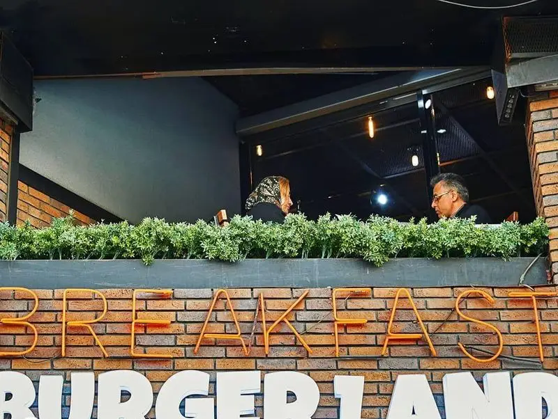 نمای بیرونی کافه رستوران برگرلند با تابلوی نئون "BREAKFAST" و "BURGERLAND"، گلدان‌های سبز و مشتریانی در حال لذت بردن از فضای داخلی.