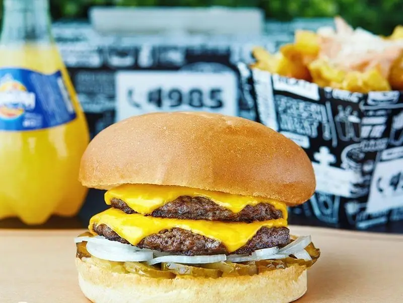 تصویری از یک دوبل چیزبرگر تازه و خوش‌طعم برگرلند با دو لایه پنیر ذوب‌شده، پیاز، خیارشور، نوشابه و سیب‌زمینی سرخ‌کرده.