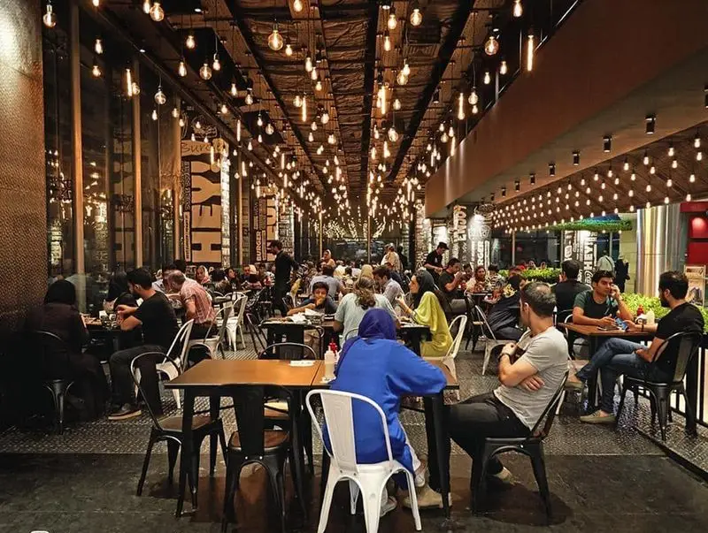 مشتریان در حال غذا خوردن در رستوران برگرلند؛ فضایی دلنشین با نورپردازی گرم و دکوراسیون خاص صنعتی و چراغ‌های آویز.