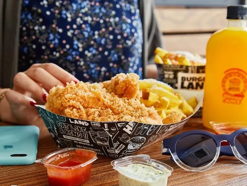 مشتری در حال لذت بردن از مرغ سوخاری ترد و سیب زمینی سرخ کرده خوشمزه برگرلند با سس‌های متنوع و آب پرتقال تازه. تجربه‌ای دلپذیر در فست فود.