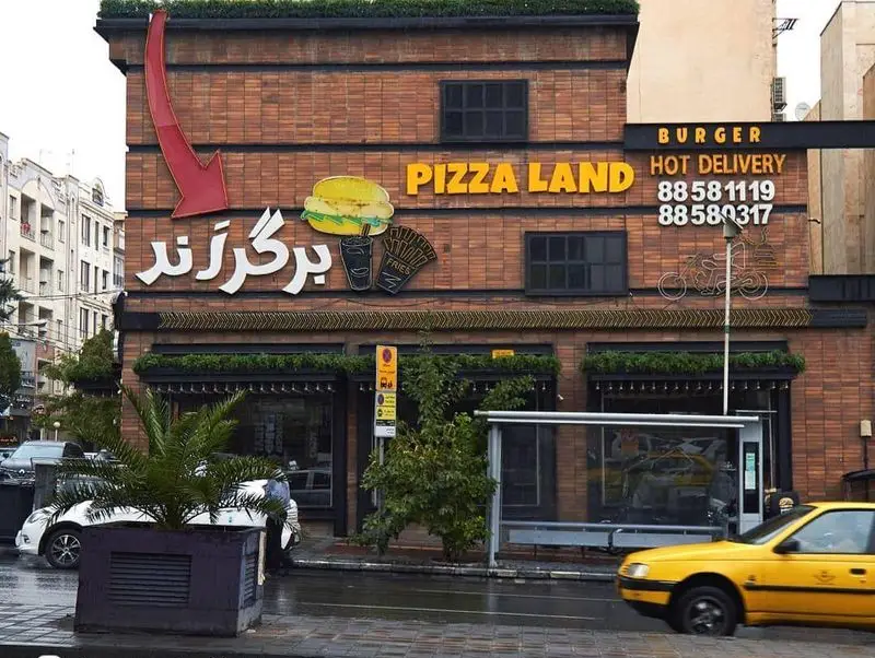 نمای بیرونی فست فود برگرلند و Pizza Land با تابلوی روشن Burger، Hot Delivery و شماره تماس، همراه با المان‌های همبرگر و موتورسیکلت تحویل.