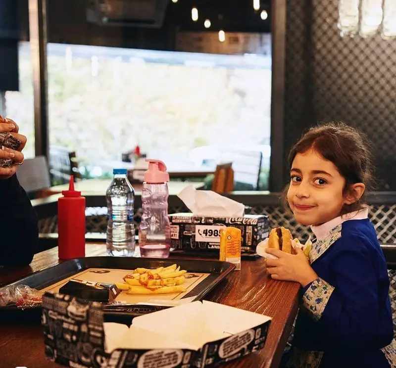 دختربچه‌ای خندان در حال لذت بردن از یک ساندویچ همبرگر خوشمزه و سیب‌زمینی سرخ کرده در فضای دنج رستوران برگرلند.