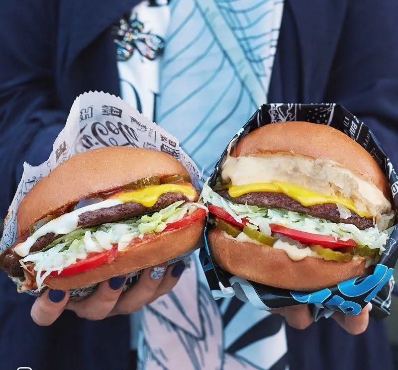 تصویر دو همبرگر لذیذ برگرلند با نان، پنیر، کاهو، گوجه و خیارشور، که در بسته‌بندی‌های مخصوص برگرلند توسط مشتریان نگه داشته شده‌اند.