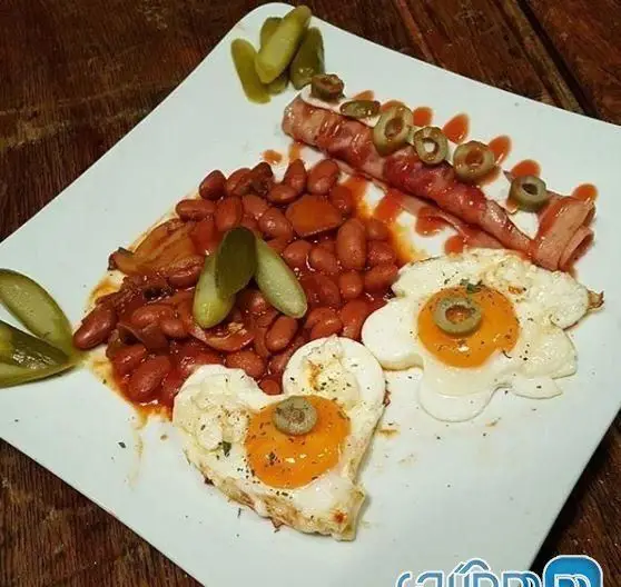 صبحانه کافه قلیان نیمکت شامل تخم مرغ های قلبی و گل، لوبیا، سوسیس بیکن و خیارشور. یک غذای خوشمزه و زیبا برای شروع روز.