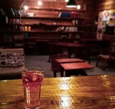 محیط دنج و گرم کافه قلیان نیمکت با کتابخانه پر از کتاب و میزهای چوبی، همراه با یک نوشیدنی خنک برای لذت بردن.