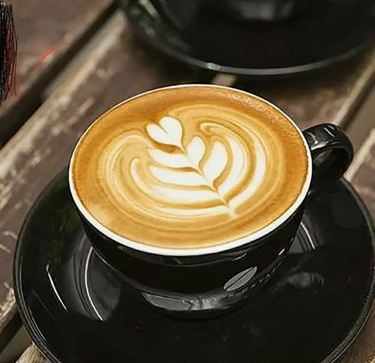 قهوه الی: فنجان مشکی لاته آرت بی‌نظیر با طرح زیبای برگ و قلب روی میز چوبی، نشان کیفیت بالای قهوه در کافه الی.