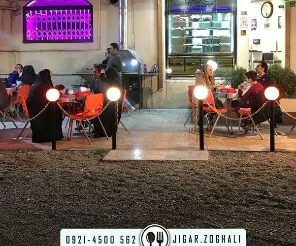 نمایی از فضای باز رستوران جیگر ذغالی با نورپردازی گرم، میز و صندلی‌های نارنجی و مشتریانی که از غذا لذت می‌برند.