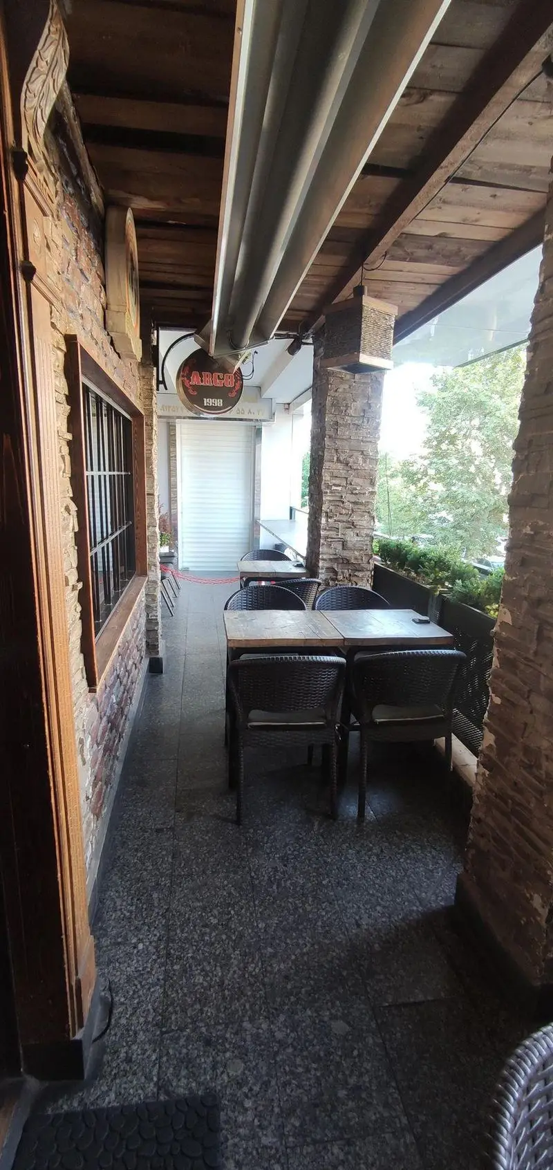 تصویر فضای باز و بالکن کافی شاپ پاییز ۹۸ با سقف چوبی، دیوارهای آجری سنگی، میز و صندلی حصیری، محیطی دنج و آرام برای مشتریان.