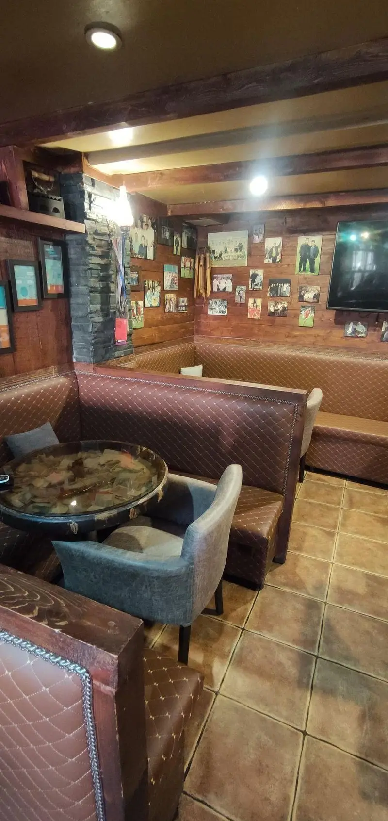 نمایی از فضای داخلی دنج و دلنشین کافی شاپ پاییز ۹۸ با نیمکت‌های چرمی کوسن‌دار، میزهای شیشه‌ای و صندلی‌های راحت، دیوارهای چوبی پر از قاب عکس.