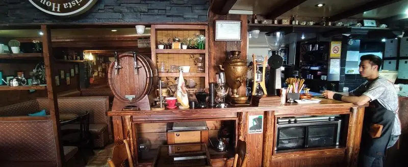 یک باریستا در کافی شاپ پاییز ۹۸، پیشخوان چوبی با انواع تجهیزات قهوه و یک بشکه چوبی زیبا، فضای داخلی دنج.