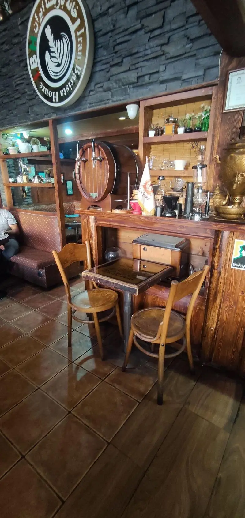 فضای داخلی دنج کافی شاپ پاییز ۹۸ با دکوراسیون چوبی، تابلو "خانه باریستا ایران"، تجهیزات متنوع قهوه و میز و صندلی‌های کلاسیک چوبی.