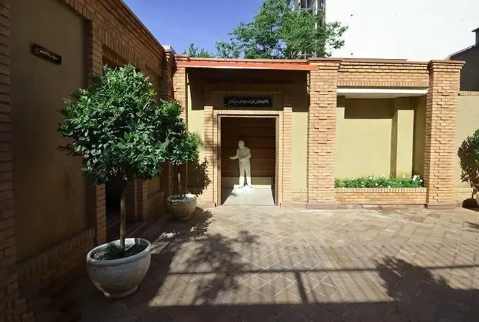 تصویری از نمای بیرونی و حیاط زیبای خانه موزه استاد عزت الله انتظامی در تهران با معماری آجری سنتی و مجسمه‌ای در ورودی مجموعه فرهنگی