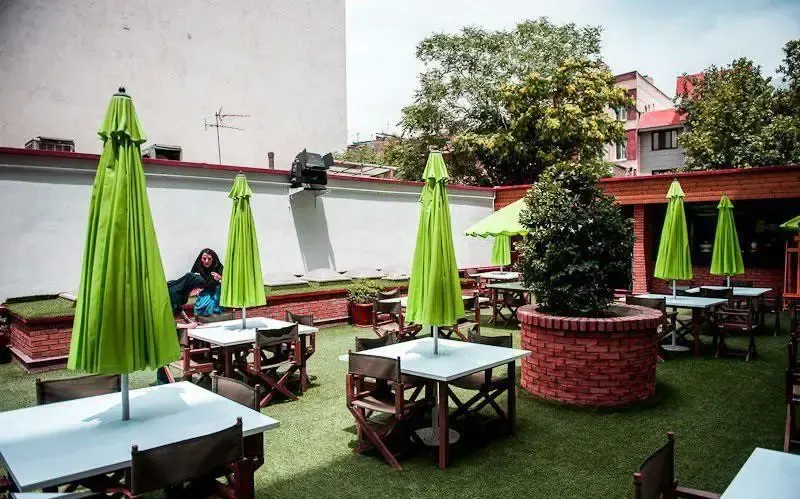 نمایی از فضای کافه روباز خانه موزه استاد عزت الله انتظامی با میز و صندلی‌های چوبی، چترهای سبز و حیاط چمن‌کاری شده.