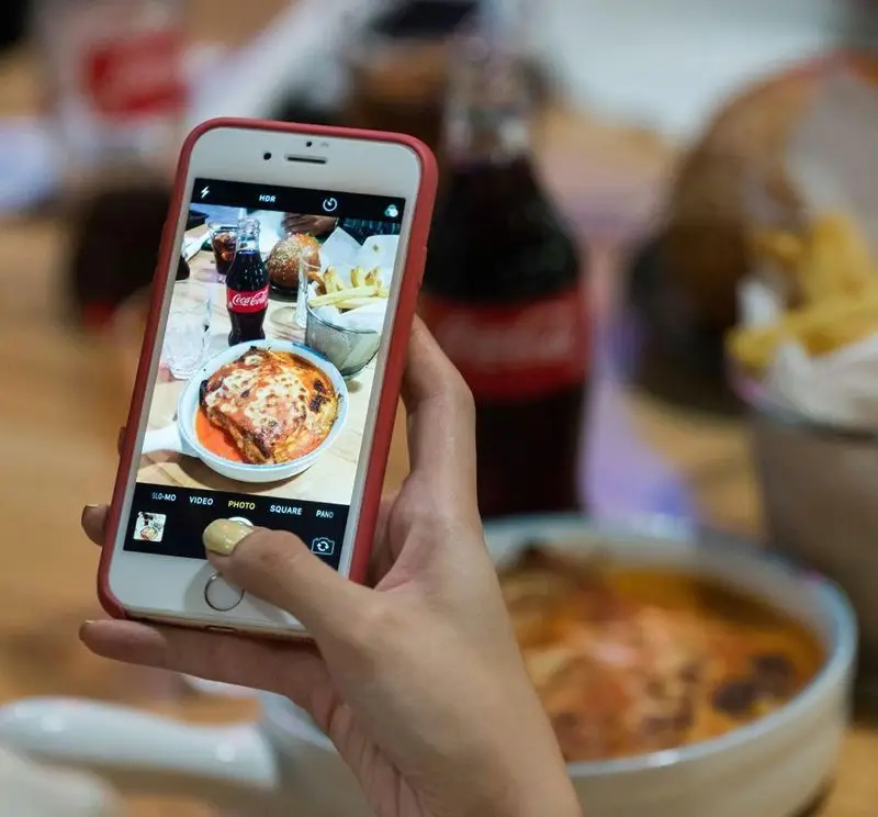 مشتری در آوافودمارکت مجتمع تجاری آواسنتر در حال عکس گرفتن از غذای خود شامل لازانیا، نوشابه و سیب زمینی سرخ کرده با گوشی.