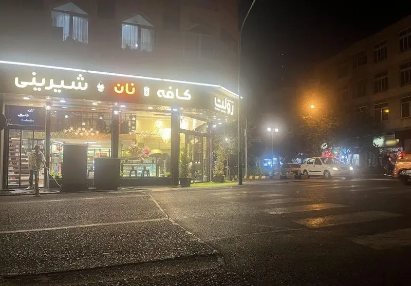 کافه قنادی رولت در شب با تابلوی نئونی کافه، نان و شیرینی. محیطی روشن و دلنشین، عابر پیاده و خودروها در خیابان شهر.