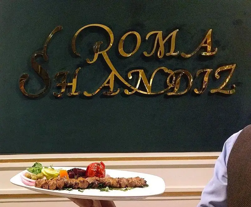 تصویر داخلی رستوران شاندیز روما با نام طلایی روی دیوار سبز و سینی کباب کوبیده با گوجه، زیتون و سبزیجات تازه توسط گارسون
