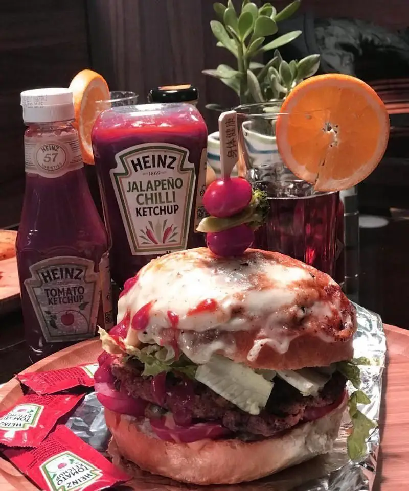 تصویری جذاب از برگر پنیری ویژه کافه رستوران فوژر، همراه با انواع سس کچاپ هاینز و یک نوشیدنی خنک؛ غذایی عالی از منوی رستوران
