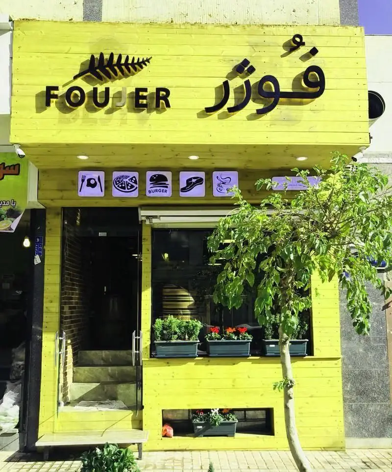 کافه رستوران فوژر: نمای بیرونی با تابلوی زرد، لوگوی برگ سرخس، و نمادهای منوی متنوع شامل صبحانه، پیتزا، برگر و نوشیدنی.