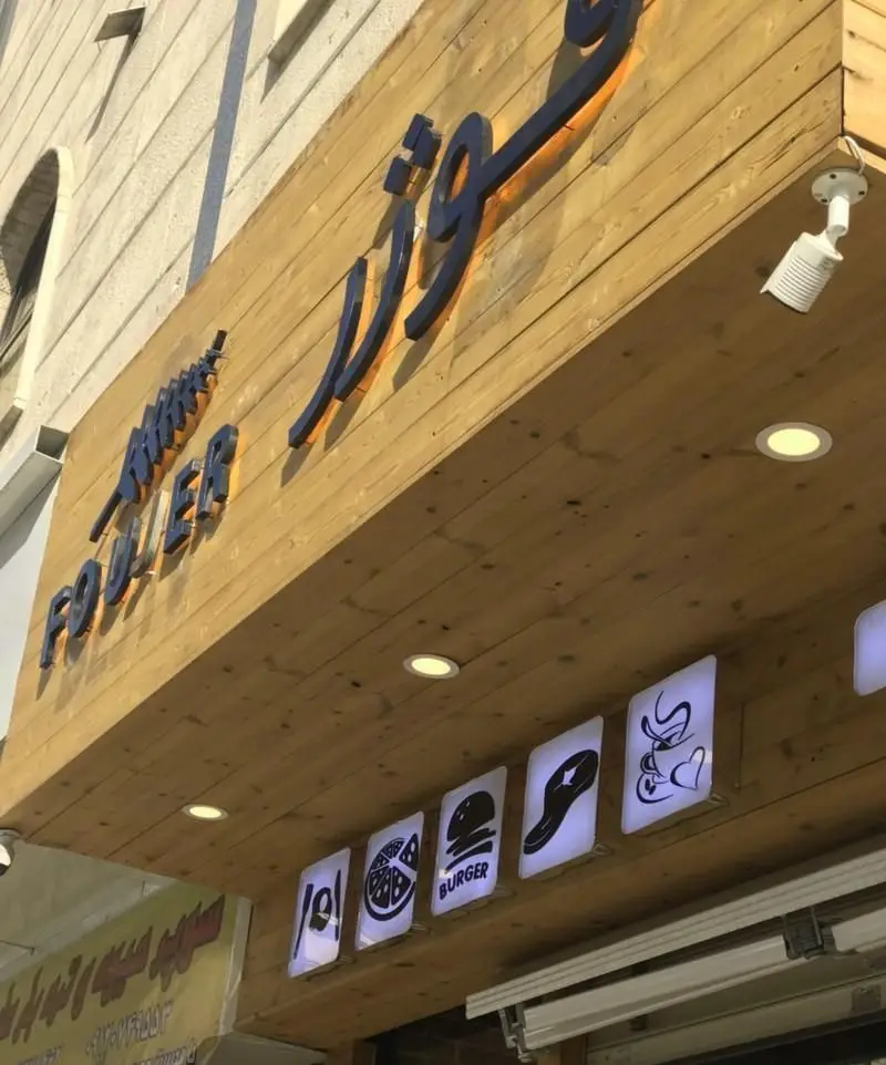 نمای بیرونی کافه رستوران فوژر با تابلوی چوبی زیبا و حروف برجسته مشکی، لوگوهای روشن غذاها شامل پیتزا، برگر، استیک و نوشیدنی گرم.