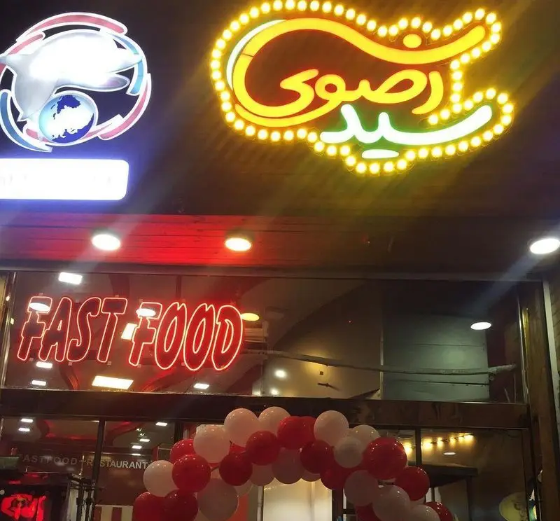 نمایی از ورودی فست فود فلفل قرمز با تابلوی نئون درخشان Fast Food، لوگو و تزئینات بادکنکی قرمز و سفید.