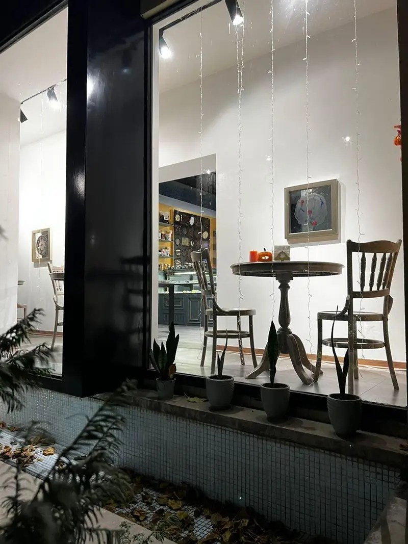 ویترین دلنشین کافه شکلات‌فروشی چوکوچاملی در شب با دکوراسیون پاییزی، شامل میز و دو صندلی چوبی، و چراغ‌های تزیینی.