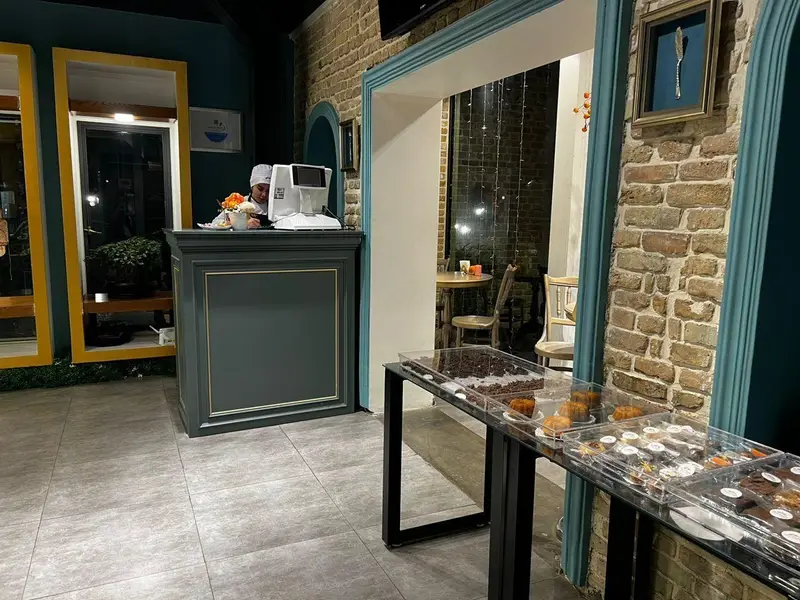 محیط داخلی زیبای کافه و شیرینی‌فروشی چوکوچاملی با پیشخوان پذیرش، دیوار آجری، میز نمایش دسرهای تازه، کیک‌ها و شیرینی‌های متنوع.