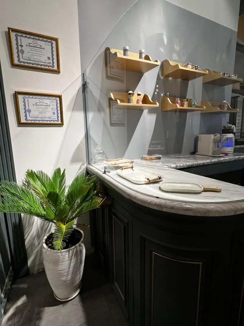 تصویر دکوراسیون داخلی کافه و شیرینی فروشی چوکوچاملی شامل پیشخوان مرمر، قفسه های چوبی و گیاه آپارتمانی سبز.